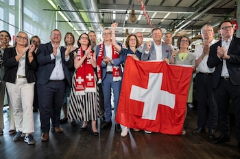 Un grupo de adultos sonrientes y aplaudiendo celebra en un interior. Varios llevan bufandas suizas rojas y blancas, y una gran bandera de Suiza se muestra
