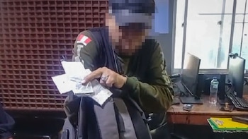 Un hombre con el rostro difuminado sostiene varios paquetes pequeños sobre una mochila en una oficina