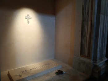Vista del sepulcro de Francisco en Roma, con la inscripción "FRANCISCVS" en la lápida, una cruz en la pared superior y ofrendas florales