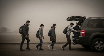 Una familia de cuatro personas con mascarillas y mochilas avanza sobre ceniza, mientras dos suben a la parte trasera de un SUV gris bajo un cielo oscuro con ceniza en el aire.