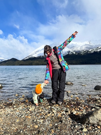 Una persona adulta y un niño pequeño caminan tomados de la mano por la orilla de un lago. Al fondo se ven montañas nevadas y un cielo azul con nubes