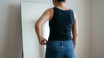Persona adulta de espaldas frente a un espejo, con musculosa negra y jeans azules holgados, separando el pantalón de su cintura con una mano.