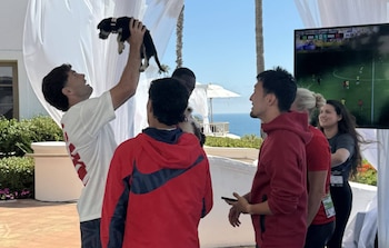 Hombre levanta cachorro. Grupo de personas. Pantalla exterior con partido de fútbol (Brasil vs Marruecos). Vista al mar, palmeras y cortinas blancas