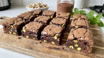 Vista cercana de una bandeja de brownies de chocolate con pistachos troceados y trozos de chocolate, sobre una tabla de madera con hojas de menta.