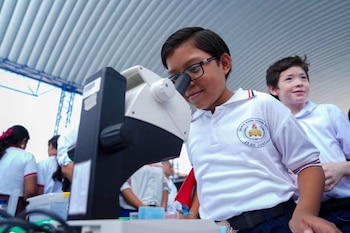 Un niño con gafas mira por un microscopio blanco y negro en una mesa, acompañado por otro alumno