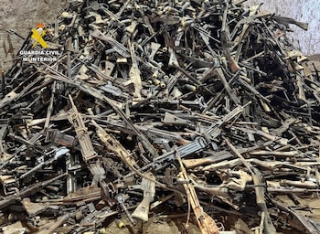 Gran pila de fusiles, armas automáticas, subfusiles y granadas apilados en un terreno, con el logo de la Guardia Civil en la esquina superior izquierda