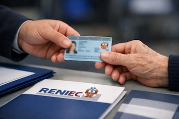 Plano medio de una mano adulta que entrega un DNI peruano azul con el escudo nacional a otra mano, sobre una mesa con folders y el logo de Reniec.