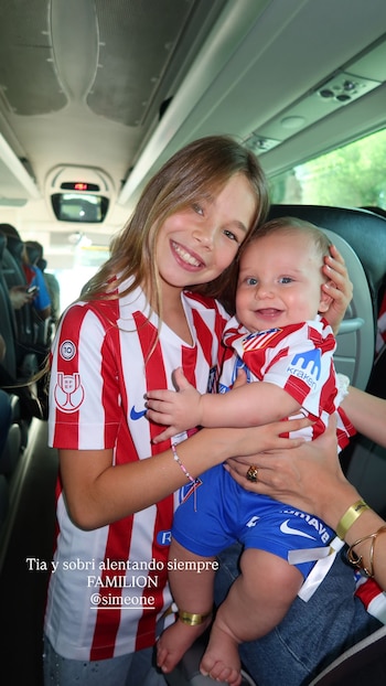Una niña con camiseta del Atlético de Madrid abraza a un bebé que también lleva la misma equipación, ambos sonriendo en el interior de un autobús