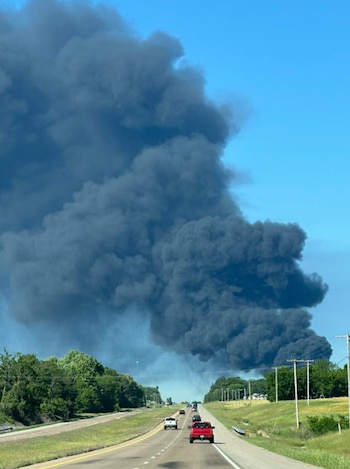 Columna masiva de humo negro y espeso se eleva hacia un cielo azul sobre una autopista con autos y árboles verdes a los lados