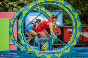 Dos mujeres en ropa deportiva, una con top rojo y otra azul, compiten en un circuito azul y amarillo con ruedas grandes y estructuras coloridas