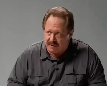 Un hombre de cabello castaño y bigote, con una camisa gris oscuro, mira hacia la derecha sobre un fondo gris liso