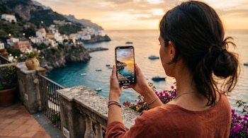 Manos de una mujer sostienen un teléfono que muestra un paisaje costero frente al mar y edificios en la montaña al atardecer.