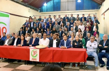 Un grupo numeroso de personas en una sala, con varios individuos sentados en una mesa roja que exhibe el logo de Juntos por el Perú, y otros de pie al fondo