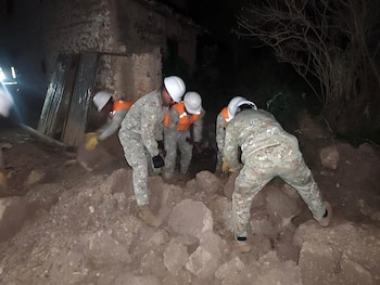 Cuatro personas con uniformes militares, chalecos reflectantes y cascos de seguridad mueven escombros en un área oscura, con una estructura dañada al fondo
