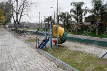 Vista exterior de un parque infantil con tobogán de colores, escaleras azules, balancines, camino de losas, césped, árboles y postes de servicios