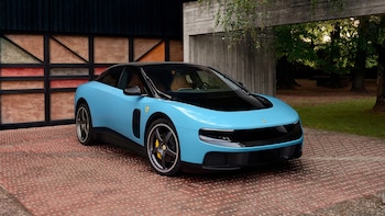 Un Ferrari Luce azul claro con techo negro y detalles en negro, estacionado en un camino de adoquines frente a un edificio moderno y árboles al fondo