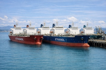 Dos buques cisterna con la palabra ETHANOL en sus costados, uno rojo y otro azul, anclados en un puerto con grúas y montañas al fondo.