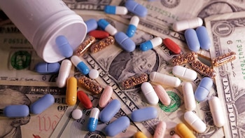 Pastillas de varios colores y cápsulas transparentes con gránulos esparcidas sobre billetes de dólar, junto a un bote blanco volcado