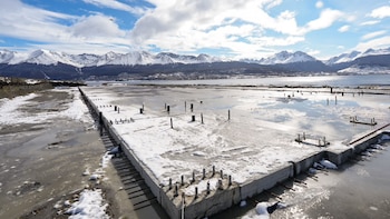 Platea de hormigón con varillas de hierro sobre un terreno con nieve y charcos de agua, con montañas nevadas de fondo