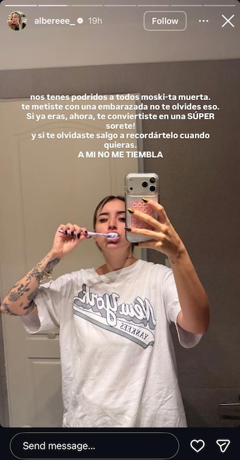Mujer joven con tatuajes y camiseta blanca se cepilla los dientes frente a un espejo, sosteniendo un teléfono con un mensaje en pantalla