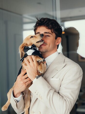 Charles Leclerc sonríe mientras sostiene a un perro pequeño de pelaje claro que lleva un esmoquin negro, el perro lo mira y nule su rostro. Leclerc viste un traje claro