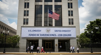 Edificio alto, bandera de Estados Unidos, pancarta blanca con texto "U.S. EMBASSY" y "Fondo de Subvenciones - USD 200,000", personas, árboles.