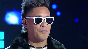 Primer plano de un hombre con gafas de sol blancas y pelo engominado, usa pendientes y collar de cuentas, contra un fondo oscuro con luces azules