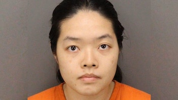 Primer plano de una mujer asiática con cabello oscuro recogido, expresión neutral, vestida con un uniforme naranja, sobre un fondo gris claro