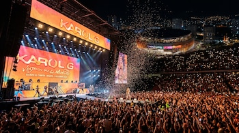 Karol G en concierto nocturno, escenario iluminado, multitud con celulares, confetti en el aire y el Arena Primavera de Sabaneta visible al fondo.