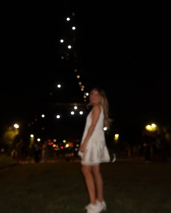 Una mujer rubia de pie, vestida con un vestido blanco y zapatillas, frente a una estructura iluminada por la noche. Fondo oscuro y borroso