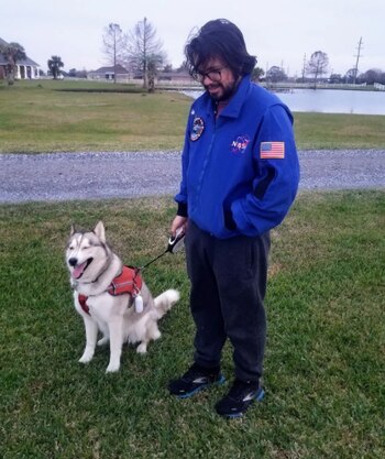Un hombre con gafas y una chaqueta azul de la NASA sonríe mientras está de pie junto a un perro husky gris y blanco con un arnés rojo, en un campo verde