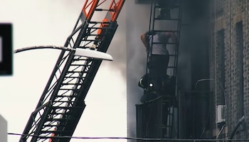 Bomberos sobre una escalera mecánica extendida hacia un edificio de ladrillo del que sale humo. Una persona y otros bomberos están en la escalera de emergencia del edificio