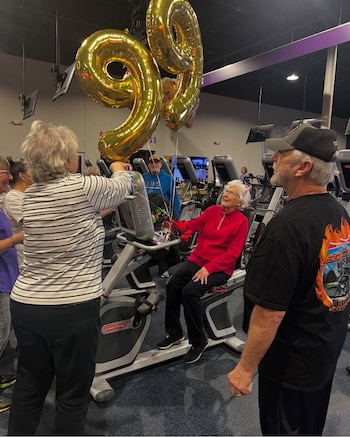 Una mujer sentada en una bicicleta fija de gimnasio rodeada de personas, con dos globos dorados con el número 99 y un ramo de flores