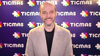 Un hombre de mediana edad con barba gris sonríe a la cámara. Viste blazer color crema y camiseta negra, frente a un muro azul con logos blancos de TICMAS
