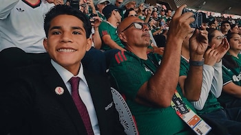 Un joven sonriente con traje oscuro y corbata burdeos posa para la cámara en un estadio de fútbol lleno de aficionados con camisetas verdes de México