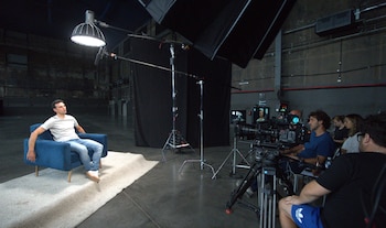 Hombre con camiseta gris y jeans en sillón azul, siendo grabado por un equipo de producción con cámaras y luces en un estudio oscuro