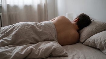 Hombre durmiendo de lado sin remera en la cama, cubierto parcialmente por un edredón claro. Hay dos almohadas, una ventana y cortinas.