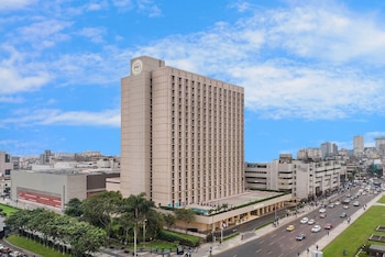 Gran edificio beige del Sheraton Lima Historic Center bajo un cielo azul con nubes, flanqueado por una avenida con coches y zonas verdes urbanas