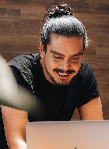Fotografía en blanco y negro de un hombre con el cabello recogido en un moño, camiseta negra de manga corta y las manos juntas