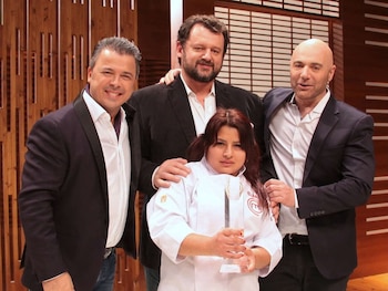 Cuatro personas posan frente a un fondo de madera. Una mujer con chaquetilla de chef blanca y trofeo. Tres hombres de traje y camisa blanca
