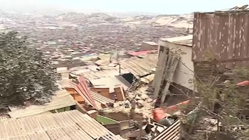 Viviendas en una ladera con techos de metal corrugado, restos de escombros de tierra y una extensa zona urbana de fondo
