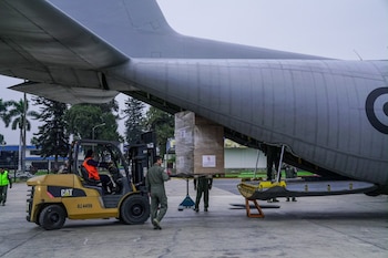 Un montacargas amarillo sube cajas de ayuda humanitaria envueltas en plástico a la rampa trasera de un avión militar gris. Se observa personal uniformado