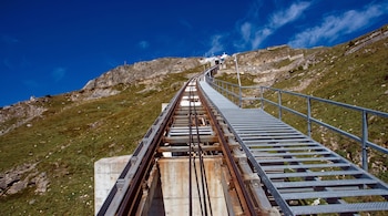 Vía de funicular con rieles metálicos, cables y cimientos de hormigón. Escalera metálica con barandilla. Ladera de montaña con rocas y pasto verde. Cielo azul.