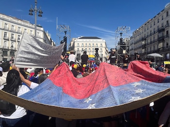 Gran bandera venezolana roja y azul con estrellas blancas, cubierta de firmas, sostenida por una multitud en una plaza con edificios y escenario al fondo