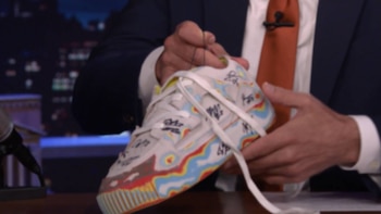 Un hombre en traje sujeta una zapatilla de deporte blanca con cordones blancos, decorada con patrones ondulados de colores rojo, azul, amarillo y negro, y escrituras en tinta negra