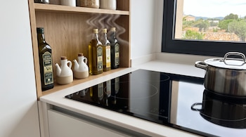 Botellas de aceite de oliva, dos aceiteras de cerámica, estante de madera, estufa de inducción negra con olla de acero inoxidable y ventana.