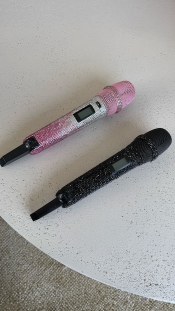 Dos micrófonos inalámbricos decorados con pedrería; uno rosa y plateado, y otro completamente negro, sobre una superficie de piedra clara
