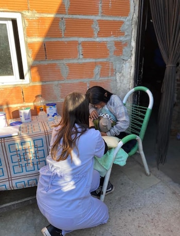 Una trabajadora de la salud con uniforme blanco se arrodilla frente a una mujer sentada en una silla, quien sostiene un bebé