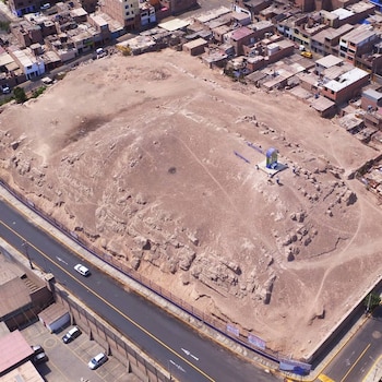 Vista aérea de la Huaca Cruz de Armatambo: extenso terreno árido con rocas. Carretera con coches y edificios residenciales alrededor. Pequeña estructura moderna y una valla visible