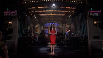 Dakota Johnson con un vestido rojo en el centro de un escenario de televisión con focos de luz y músicos al fondo